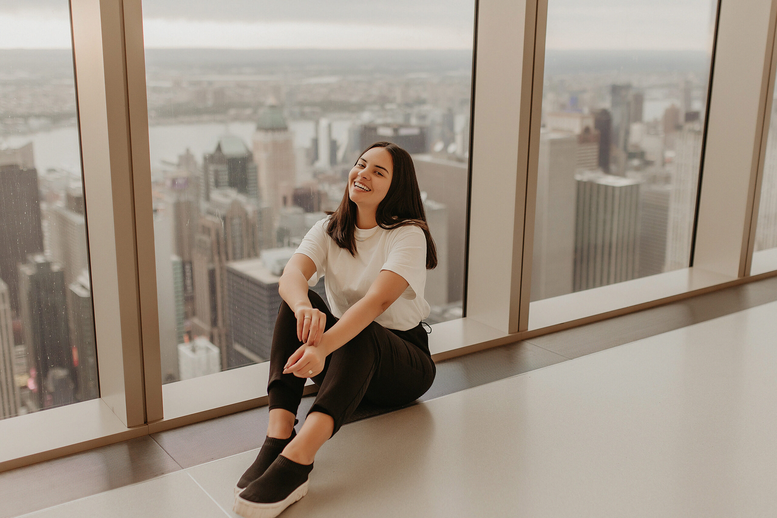 Isis Castro sentada no topo do Summit, observatório em NY. Ela está sentada no chão, perto da janela e sorrindo, representando uma das conquistas de ser uma fotógrafa de sucesso.