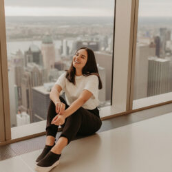 Isis Castro sentada no topo do Summit, observatório em NY. Ela está sentada no chão, perto da janela e sorrindo, representando uma das conquistas de ser uma fotógrafa de sucesso.