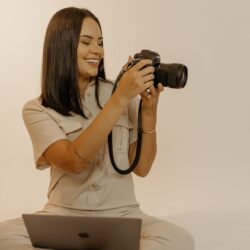 Isis está com seu notebook no colo e sua câmera fotografando na mão. Esta é uma foto para o lançamento do curso de fotografia online MOVE.